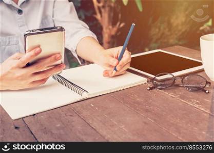 asian woman hand holding phone and pencil writing notebook in coffee shop with vintage toned