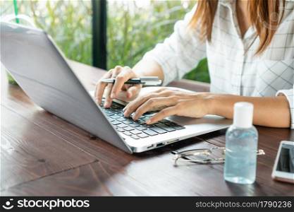 Asian woman hands working on computer with sanitizer gel; mobile phone and glasses on table. Protective measures to prevent COVID-19 transmission; bisiness and health care concept. Close up