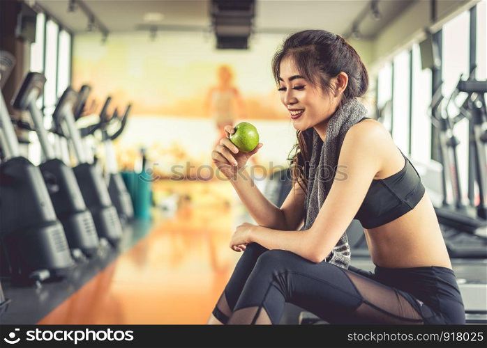 Asian woman holding and looking green apple to eat with sports equipment and treadmill in background. Clean food and Healthy concept. Fitness workout and running theme.