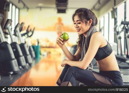 Asian woman holding and looking green apple to eat with sports equipment and treadmill in background. Clean food and Healthy concept. Fitness workout and running theme.
