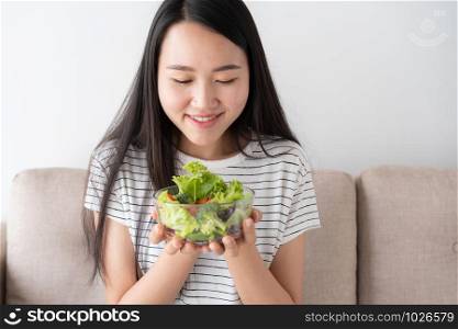 Asian woman holds a glass bowl full of salad vegetables green while sitting on the sofa at home. healthy and lifestyle concept