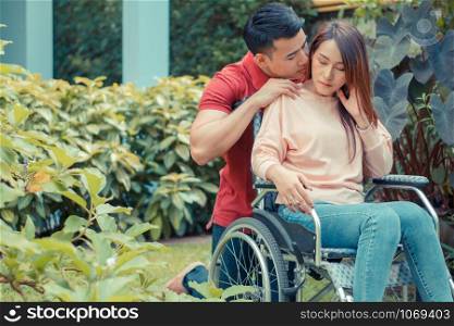 Asian woman in a wheelchair and Unhappy and painful. A Man standing behind the wheelchair and is encouraging his wife, whose feet hurt her leg due to an accident. Concept of caring and support