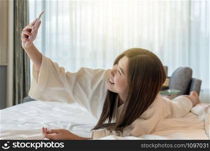 Asian woman in bathrope suit using the smart mobile phone with smiling action to selfie herself on the bed when wake up in luxury hotel, lifestyle and leisure concept