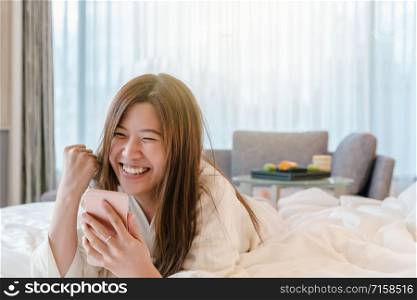 Asian woman in bathrope suit using the smart mobile phone with success action on the bed when wake up in luxury hotel, lifestyle and leisure concept