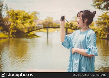 Asian woman in blue dress in public park carrying digital mirrorless camera and taking photo without facial mask in happy mood. People lifestyle and leisure concept. Outdoor travel and Nature theme.