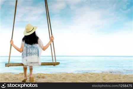 Asian woman in casual style wear hat and sandals sit on swings at sand beach and looking beautiful tropical paradise sea and sky. Summer vacation. Summer vibes. Enjoying and relaxing girl on holiday.