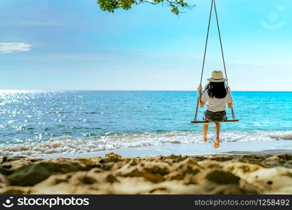 Asian woman in casual style wear hat swing the swings at sand beach and looking beautiful tropical paradise sea and sky on sunset. Summer vacation. Summer vibes. Enjoying and relaxing girl on holiday