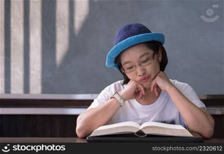 Asian woman in eyeglasses prop up on chin with suspicious and tired expression on her face while reading textbook in vintage living room