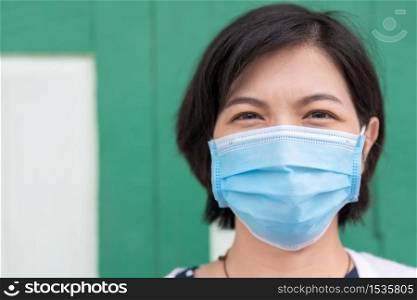Asian woman in face mask over green background looking at camera at outdoor. Young female with new normal and social distancing during pandemic coronavirus or covid-19.