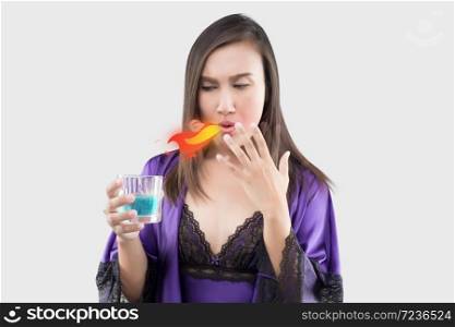 Asian woman in sexy silk nightgown and purple robe feel burn in her mouth because using mouthwash against a gray background, Wash the mouth, Female rinsing and gargling while using mouthwash from a glass