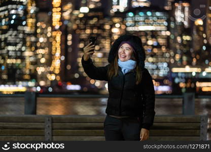 Asian woman in Winter suit using smart mobile phone for selfie over the photo blurred bokeh of New york Cityscape beside the east river background, USA downtown, career concept