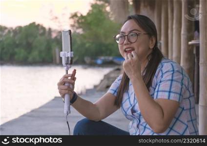 Asian woman is applying lipstick with using mobile phone instead of a mirror while relaxing on sea viewpoint at sunset time