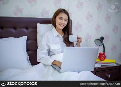 Asian woman is drinking a cup of coffee and using a notebook to work on the bed.She smiles and enjoys working at home.