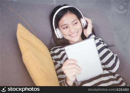 Asian woman is resting, listening to music on sofa with earphones.
