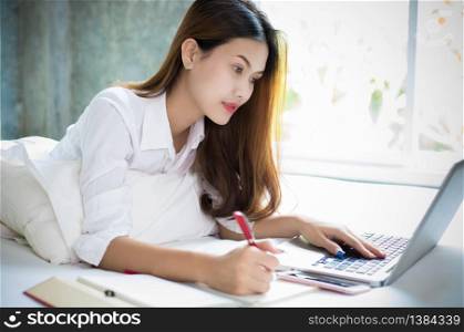 Asian woman is using a pen to write notes in her notebook and she uses a notebook to work on her bed at home.