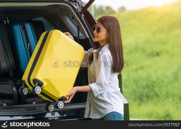 Asian woman lifting yellow suitcase into SUV car during travel in long weekend trip. People lifestyles and transportation concept. Girl put luggage from car trunk to camping. Selective focus on car