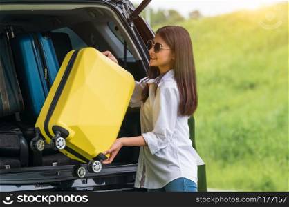 Asian woman lifting yellow suitcase into SUV car during travel in long weekend trip. People lifestyles and transportation concept. Girl put luggage from car trunk to camping. Selective focus on car