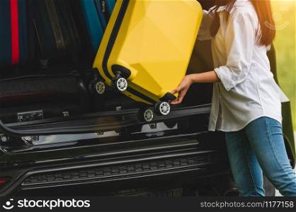 Asian woman lifting yellow suitcase into SUV car during travel in long weekend trip. People lifestyles and transportation concept. Girl put luggage from car trunk to vacation camping in summer time