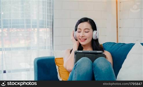 Asian woman listening music and using tablet, female using relax time lying on home sofa in living room at home. Happy female listening music with headphones concept.
