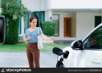 Asian woman lock electric vehicle with remote car key at her garage, EV car recharge battery at home charging station. Alternative clean energy applied in daily life as progressive lifestyle concept.. Progressive concept of asian woman and electric car with home charging station.