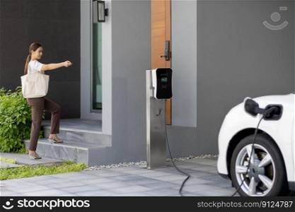 Asian woman lock electric vehicle with remote car key at her garage, EV car recharge battery at home charging station. Alternative clean energy applied in daily life as progressive lifestyle concept.. Progressive concept of asian woman and electric car with home charging station.