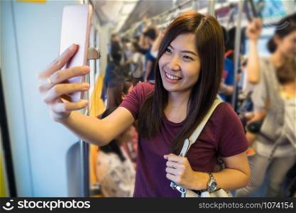 Asian woman passenger with casual suit taking selfie by the smart mobile phone in the Skytrain rails or subway for travel in the big city, lifestyle and transportation concept
