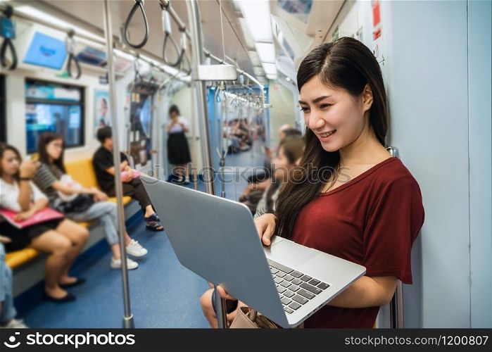 Asian woman passenger with casual suit using the technology laptop in the BTS Skytrain rails or MRT subway for travel in the big city, lifestyle and transportation concept
