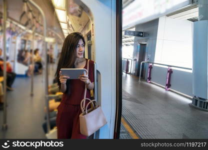 Asian woman passenger with casual suit using the technology tablet in the BTS Skytrain rails or MRT subway for travel in the big city, lifestyle and transportation concept