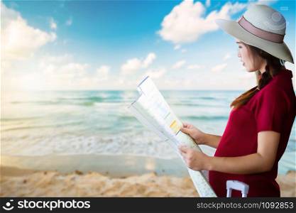 Asian woman pregnant in red dress carrying red luggage and look at the map at early morning sunrise over and Wave of the sea on the sand beach the horizon background,Holiday travel concept.