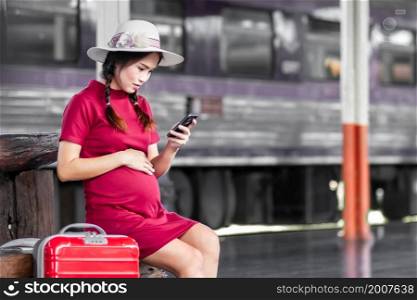 Asian woman pregnant in red dress carrying red luggage and look at the Smartphone with a red suitcase at railway station travel,traveler with backpack in summer Holiday concept Thailand