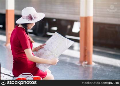 Asian woman pregnant in red dress carrying red luggage and look at the map at railway station travel.