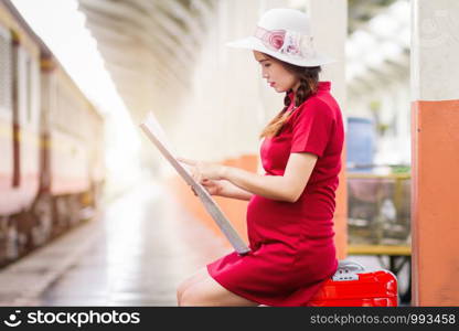 Asian woman pregnant in red dress carrying red luggage and look at the map at railway station travel.