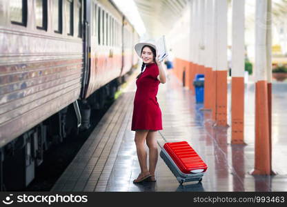 Asian woman pregnant in red dress holding a map and hand up say hello with red suitcase in railway station travel.