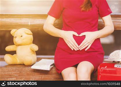 Asian woman pregnant red dress Heart-shaped handmade sitting on a bench with Teddy Bear and map with red suitcase at railway station travel.