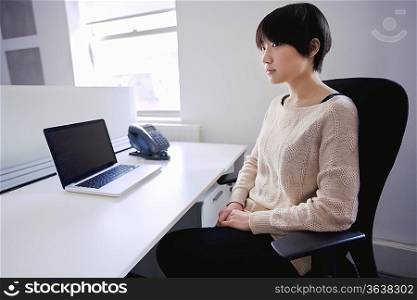 Asian woman sitting at desk with laptop while looking away