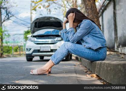 Asian woman sitting beside car after a car breakdown on street. Concept of vehicle engine problem or accident and emergency help from Professional mechanic