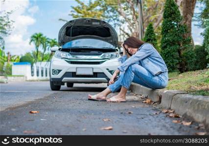 Asian woman sitting beside car after a car breakdown on street. Concept of vehicle engine problem or accident and emergency help from Professional mechanic