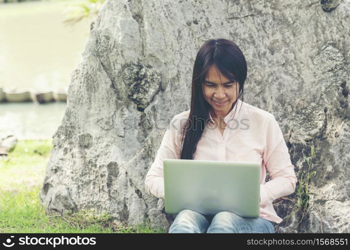 Asian woman sitting green park using laptop computer. Woman working on laptop happy entrepreneur business using notebook with hands typing on keyboard home office during coronavirus quarantine period