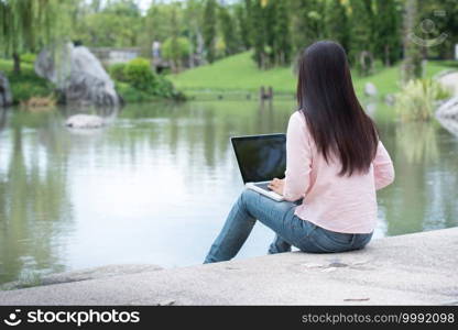 Asian woman sitting green park using laptop computer. Woman working on laptop happy entrepreneur business using notebook with hands typing on keyboard home office during coronavirus quarantine period