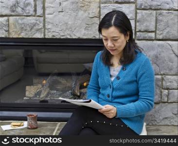 Asian woman sitting next to fireplace reading magazine with coffee cup next to her