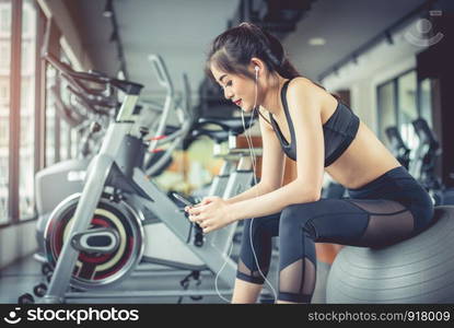 Asian woman sitting on fitness ball and listening music in fitness training gym with smart phone. Relax and sport workout training concept. Technology and Entertainment theme