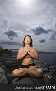 Asian woman sitting on rock by ocean in lotus pose with eyes closed in Maui, Hawaii