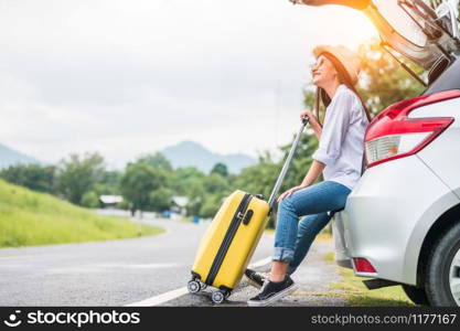 Asian woman spending weekend in roadtrip with yellow luggage. Girl relaxing on back of car with road background. Road trip and holiday vacation concept. People lifestyles and transportation.