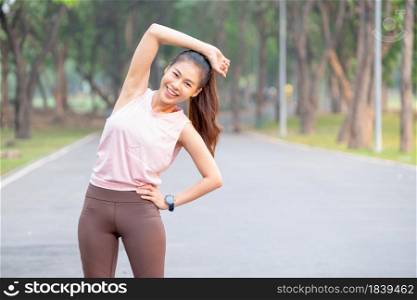 Asian woman stand and do right arm stretching also look at camera and smile in park or garden with road and tree as background and morning light.