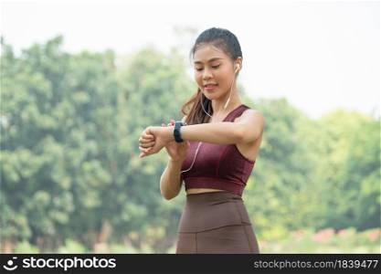 Asian woman stand and look at her watch during exercise in park or garden with green tree and sky as background.