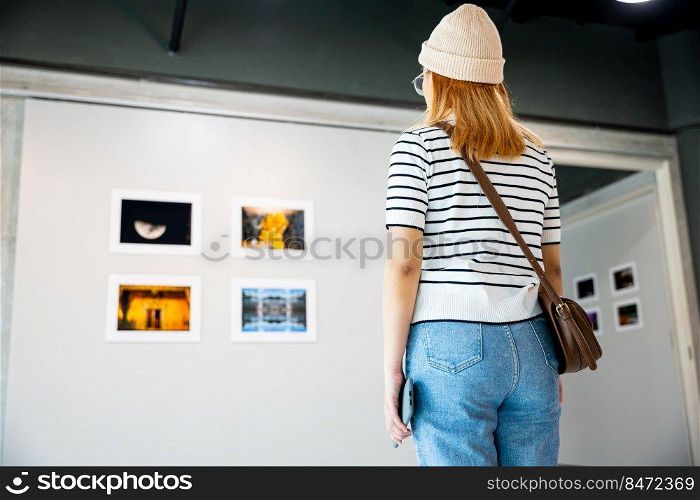 Asian woman standing she looking art gallery in front of colorful framed paintings pictures on white wall, people watch at photo frame to leaning against at show exhibition artwork gallery, Back view
