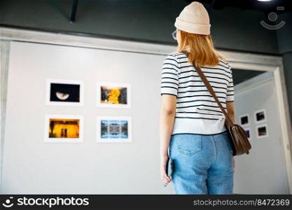 Asian woman standing she looking art gallery in front of colorful framed paintings pictures on white wall, people watch at photo frame to leaning against at show exhibition artwork gallery, Back view