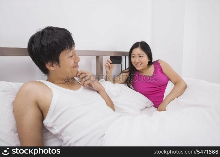 Asian woman taking a picture of man with cell phone in bed