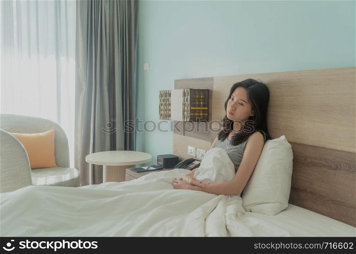 Asian woman thinking about problems and suffering from depression on bed in a modern bedroom with white blanket.