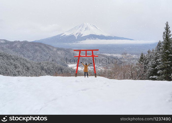 Asian woman tourist travel at Red Japanese Torii pole, Fuji mountain and snow in Kawaguchiko, Japan. Forest trees nature landscape background in winter season.